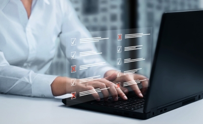 Attēlā redzams cilvēks, kurš raksta uz klēpjdatora tastatūras, bet virs tā redzama caurspīdīga digitāla anketa ar atzīmju rūtiņām, kurās daļa atbilžu ir apstiprinātas ar ķeksīšiem, bet citas noraidītas ar sarkaniem krustiņiem.