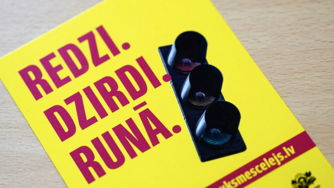 Dzeltens informatīvs plakāts ar uzrakstu “REDZI. DZIRDI. RUNĀ.” un luksofora attēlu; apakšā redzama tīmekļa adrese “traukescelejs.lv”.