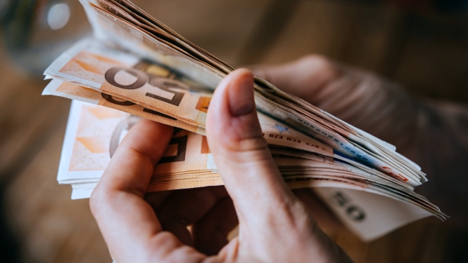 Attēlā redzamas personas rokas, kurās tiek turētas eiro banknotes. 