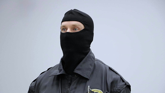 Attēlā redzama persona ar melnu seju aizsedzošu masku un virsjakā, uz kuras redzams KNAB logo