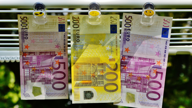 Trīs eiro banknotes – divas 500 eiro un viena 200 eiro – piestiprinātas ar knaģiem pie veļas auklas ārā, it kā žūstot; fonā izplūdis zaļš dārza skats.