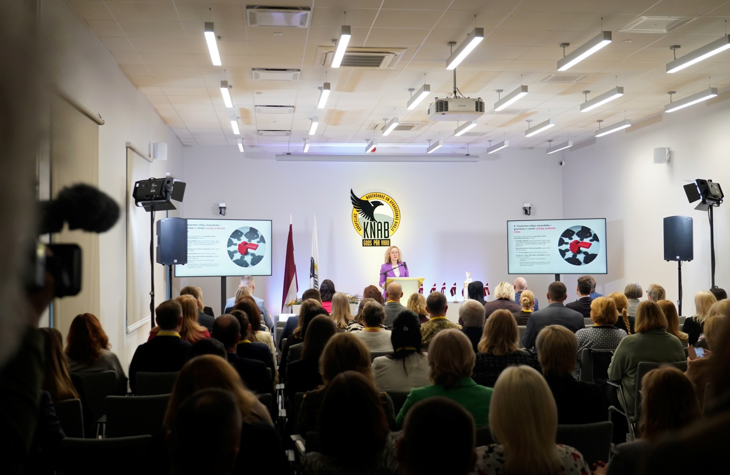 Attēlā redzama KNAB Konferenču zāle pilna ar cilvēkiem