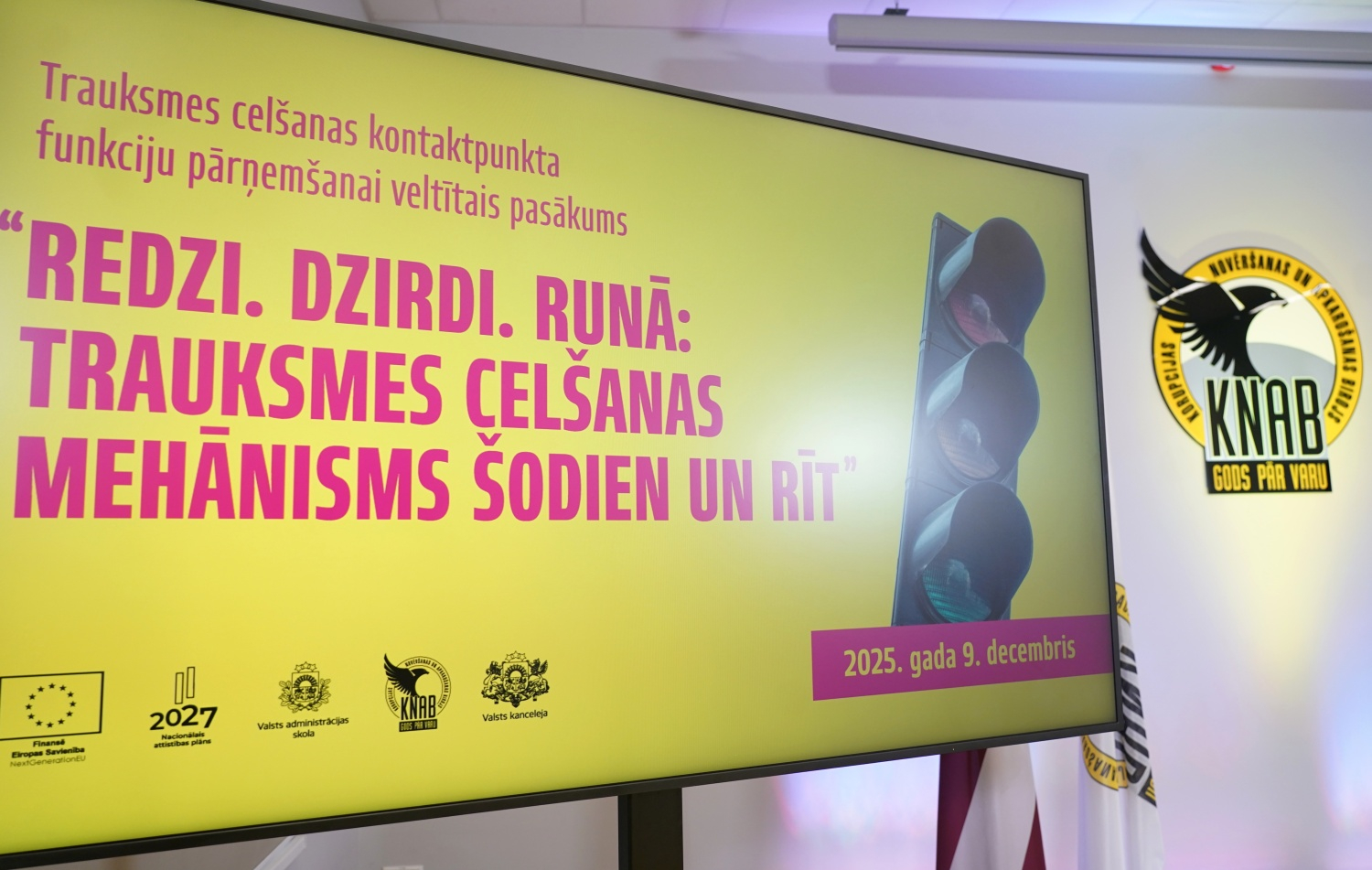 Attēlā redzams liels prezentācijas ekrāns ar dzeltenu fonu, uz kura latviski rakstīts pasākuma nosaukums “Redzi. Dzirdi. Runā: trauksmes celšanas mehānisms šodien un rīt”. Labajā pusē redzams attēls ar luksoforu, bet fonā – Korupcijas novēršanas un apkarošanas biroja emblēma un Latvijas karogs. Ekrāna apakšā atrodami organizatoru un ES fondu logo, kā arī norādīts datums “2025. gada 9. decembris”.