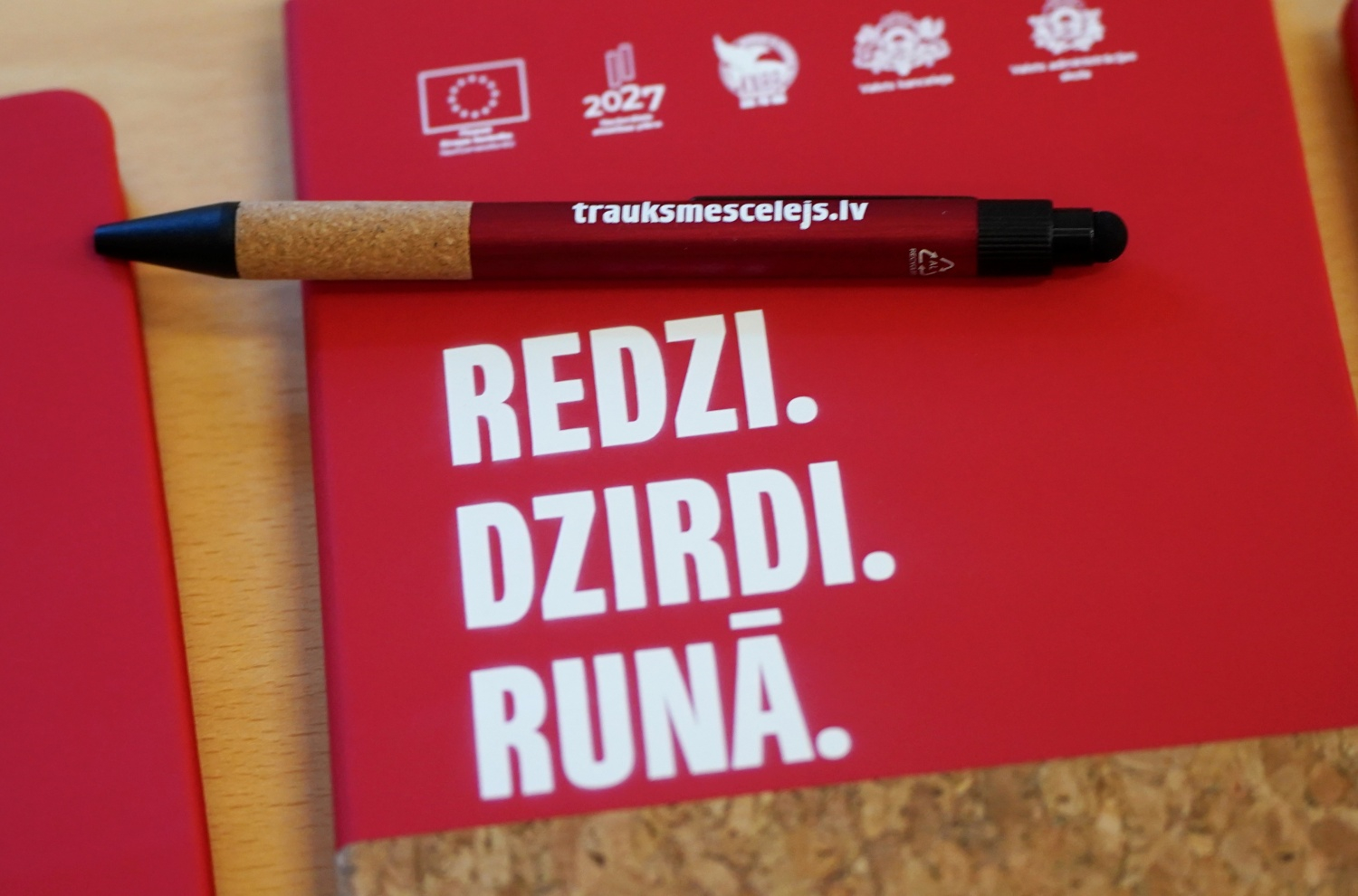 Attēlā redzama pildspalva ar uzrakstu "trauksmescelejs.lv" un piezīmju bloknots ar uzrakstu "Redzi. Dzirdi.Runā"