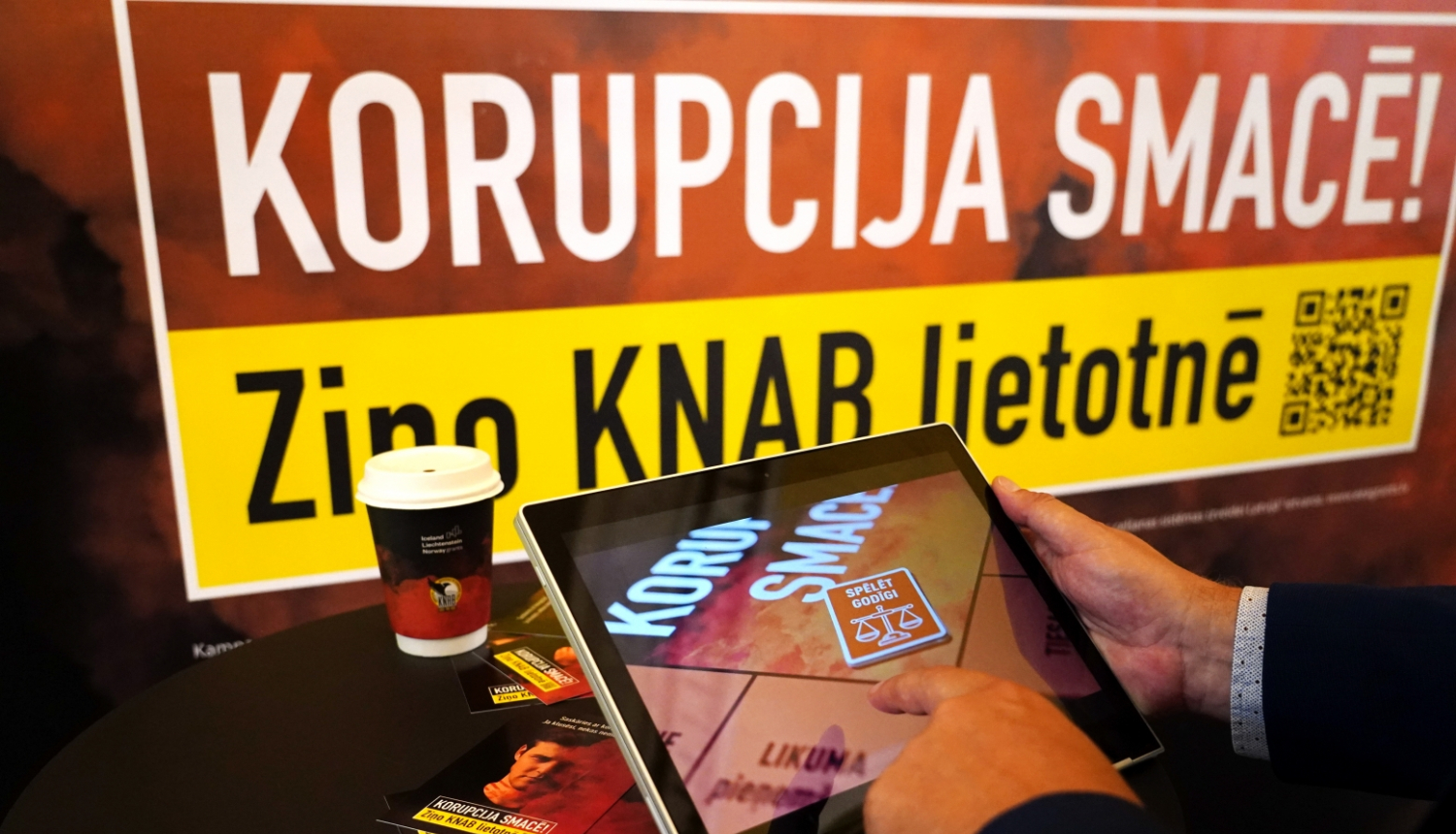 Baneris ar kampaņas devīzi "Korupcija smacē! Ziņo KNAB lietotnē", kā arī attēls ar planšetdatoru, kurā spēlēts kampaņas interaktīvais rīks