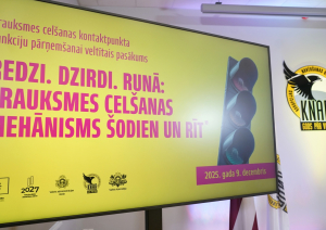 Attēlā redzams liels prezentācijas ekrāns ar dzeltenu fonu, uz kura latviski rakstīts pasākuma nosaukums “Redzi. Dzirdi. Runā: trauksmes celšanas mehānisms šodien un rīt”. Labajā pusē redzams attēls ar luksoforu, bet fonā – Korupcijas novēršanas un apkarošanas biroja emblēma un Latvijas karogs. Ekrāna apakšā atrodami organizatoru un ES fondu logo, kā arī norādīts datums “2025. gada 9. decembris”.