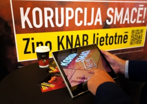 Baneris ar kampaņas devīzi "Korupcija smacē! Ziņo KNAB lietotnē", kā arī attēls ar planšetdatoru, kurā spēlēts kampaņas interaktīvais rīks