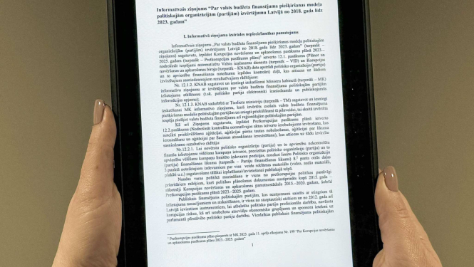 Attēlā redzama planšete, kuru tur rokās un uz kuras redzams teksts no informatīvā ziņojuma projekta, kas saistīts ar valsts budžeta finansējuma piešķiršanas modeļa politiskajām organizācijām (partijām) izvērtējumu Latvijā no 2018. gada līdz 2023. gadam.