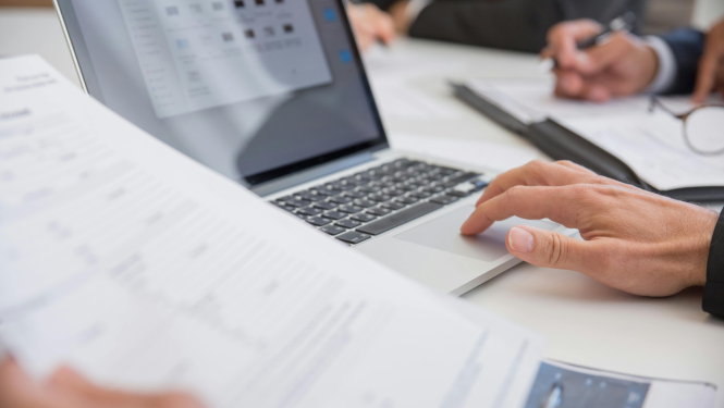Attēlā redzamas cilvēka rokas pie datora, vienlaikus pārskatot dokumentus. Fonā redzams kolēģis, kas pieraksta piezīmes uz papīra