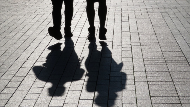 Attēlā redzami divu cilvēku silueti, tie redzami no ceļgaliem uz leju, ejot pa bruģētu ietvi; uz zemes skaidri redzamas viņu garās ēnas.