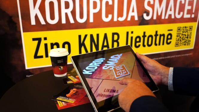 Baneris ar kampaņas devīzi "Korupcija smacē! Ziņo KNAB lietotnē", kā arī attēls ar planšetdatoru, kurā spēlēts kampaņas interaktīvais rīks