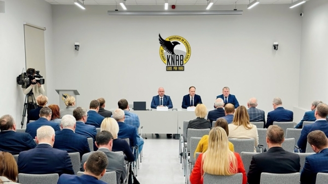 Gaiša zāle, fonā KNAB logo. Telpā ar muguru sēž dažādi cilvēki, zāles galā pie galda trīs vīrieši. No kreisās: KNAB priekšnieks Jēkabs Straume, Eiropas prokurors Gatis Doniks un Latvijas pašvaldību savienības priekšsēdis Gints Kaminskis. 