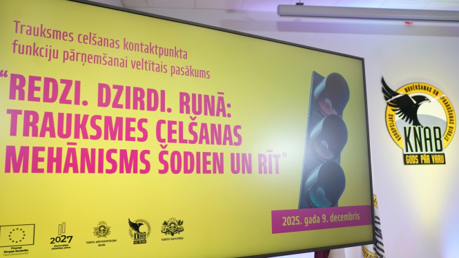 Attēlā redzams liels prezentācijas ekrāns ar dzeltenu fonu, uz kura latviski rakstīts pasākuma nosaukums “Redzi. Dzirdi. Runā: trauksmes celšanas mehānisms šodien un rīt”. Labajā pusē redzams attēls ar luksoforu, bet fonā – Korupcijas novēršanas un apkarošanas biroja emblēma un Latvijas karogs. Ekrāna apakšā atrodami organizatoru un ES fondu logo, kā arī norādīts datums “2025. gada 9. decembris”.