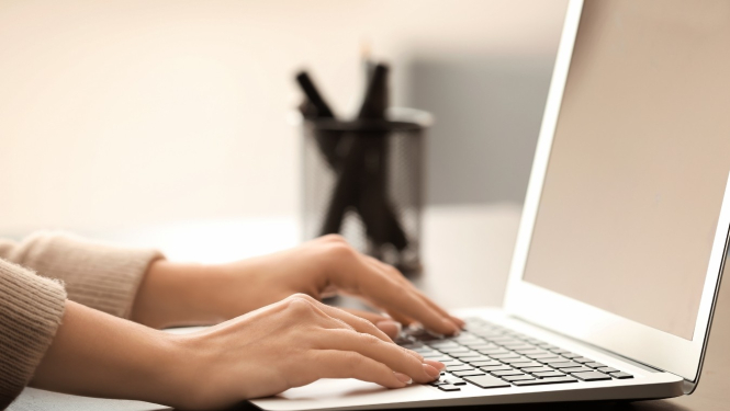 Attēlā redzamas rokas, kas raksta uz portatīvā datora tastatūras. Uz galda blakus datoram atrodas arī viedtālrunis un pildspalvu turētājs ar pildspalvām.