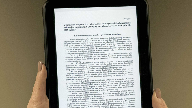 Attēlā redzama planšete, kuru tur rokās un uz kuras redzams teksts no informatīvā ziņojuma projekta, kas saistīts ar valsts budžeta finansējuma piešķiršanas modeļa politiskajām organizācijām (partijām) izvērtējumu Latvijā no 2018. gada līdz 2023. gadam.