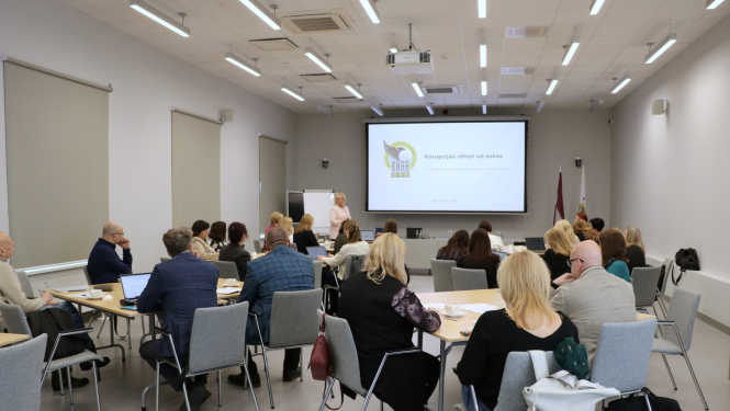 Cilvēku grupa sēž konferenču zālē pie vairākiem galdiem un klausās prezentāciju. Priekšā stāv sieviete, kas vada apmācību, aiz viņas uz ekrāna redzama prezentācijas slaids ar uzrakstu “Korupcijas cēloņi un sekas” un KNAB logo. Telpā ir gaišas sienas, lieli logi ar aizritinātiem aizkariem un griestos iebūvēti apgaismojuma paneļi. Auditorijā redzami cilvēki ar klēpjdatoriem un dokumentiem.