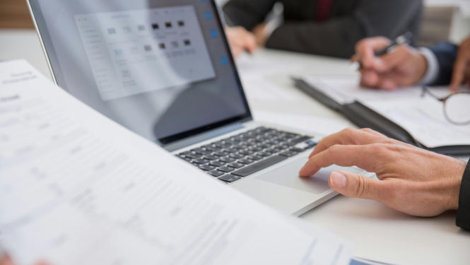 Attēlā redzamas cilvēka rokas pie datora, vienlaikus pārskatot dokumentus. Fonā redzams kolēģis, kas pieraksta piezīmes uz papīra