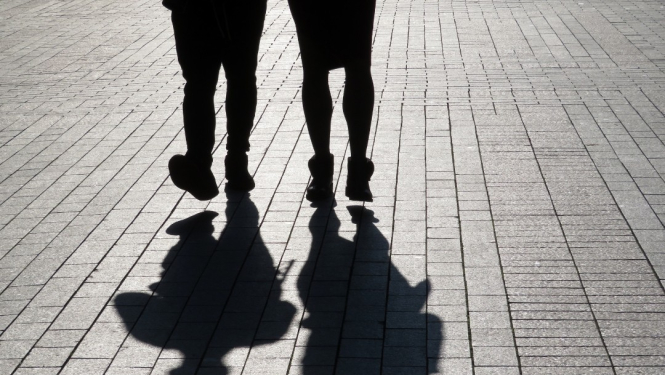 Attēlā redzami divu cilvēku silueti, tie redzami no ceļgaliem uz leju, ejot pa bruģētu ietvi; uz zemes skaidri redzamas viņu garās ēnas.