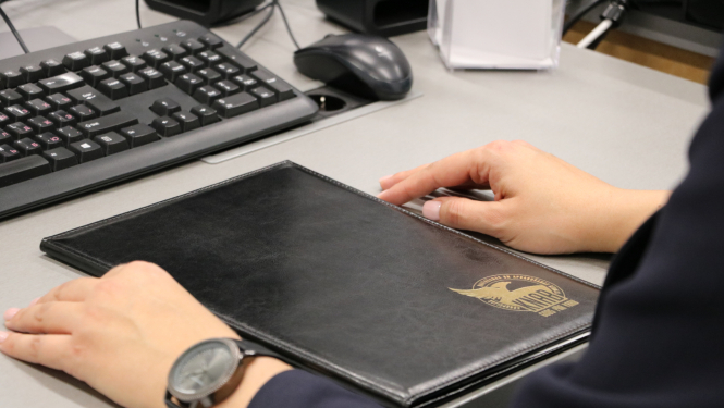 Attēlā redzams galds, uz kura stāv klaviatūra, mape ar KNAB logo un persona novietojusi rokas