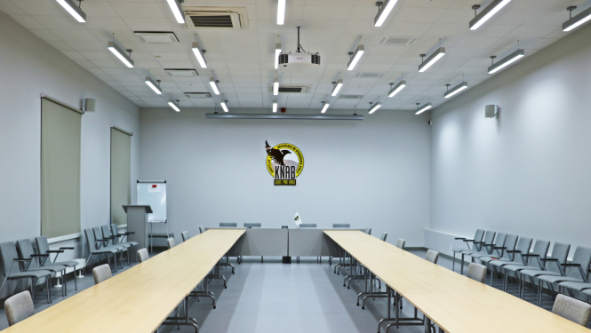KNAB zāle. Gaiša telpa, tās galā pie sienas KNAB logo. Izkārtoti divi garie galdi rindās, pie kurām novietoti krēsli
