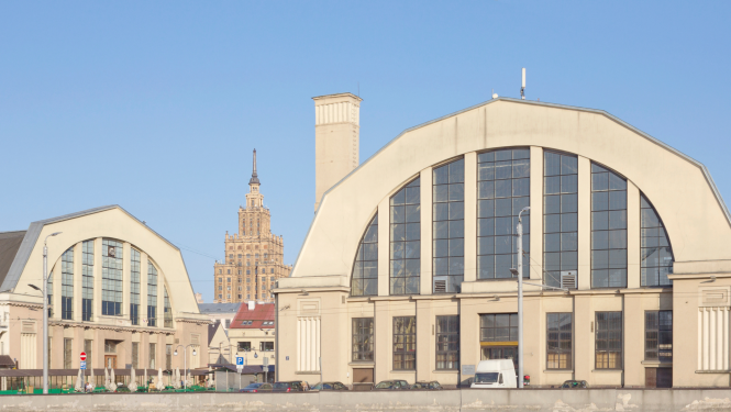 Rīgas Centrāltirgus paviljoni ar lieliem pusapaļiem jumtiem un stiklotām fasādēm saulainā dienā; fonā redzama Latvijas Zinātņu akadēmijas ēka un zilas debesis.