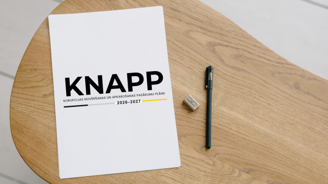Uz koka galda novietota balta brošūra ar nosaukumu 'KNAPP – Korupcijas novēršanas un apkarošanas pasākumu plāns 2026–2027'. Blakus redzama melna pildspalva un asināmais