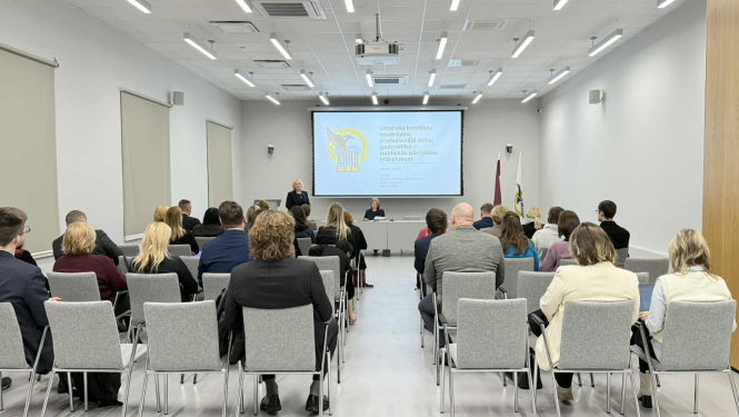 Attēlā redzama KNAB konferenču zāle, kurā notiek mācības par interešu konflikta novēršanu, profesionālo ētiku valsts pārvaldē. Auditorijā sēž klausītāji, priekš pie galda runā divas KNAB pārstāves. Uz ekrāna redzama prezentācija ar KNAB logo un temata nosaukumu. Tāpat telpā redzams Latvijas valsts karogs un KNAB karogs. 