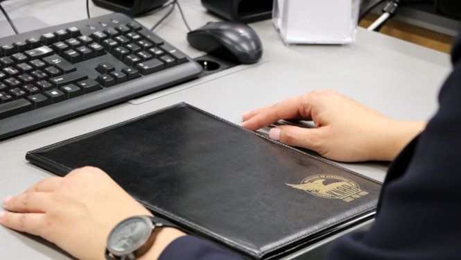 Persona sēž pie biroja galda ar klaviatūru un peli; uz galda priekšā atrodas aizvērta melna ādas mape ar zeltītu KNAB logotipu, redzamas personas rokas un rokas pulkstenis