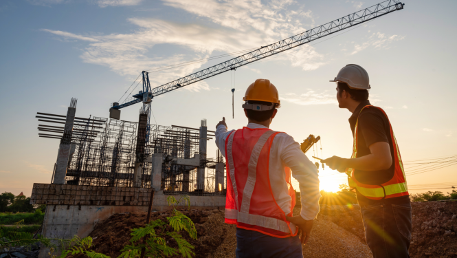 Divi būvnieki aizsargķiverēs un drošības vestēs apskata būvlaukumu saulrietā, viens rāda uz daļēji uzbūvētu konstrukciju un celtniecības celtni fonā.