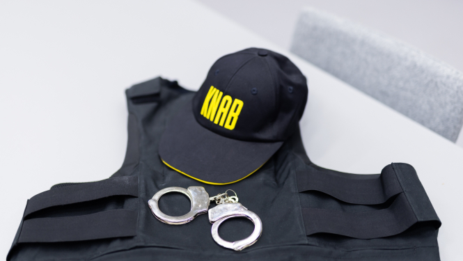 Melna aizsargvestes tipa veste ar uz tās novietotu melnu cepuri ar dzeltenu uzrakstu “KNAB” un sudraba krāsas roku dzelžiem, kas atrodas uz gaiša galda.