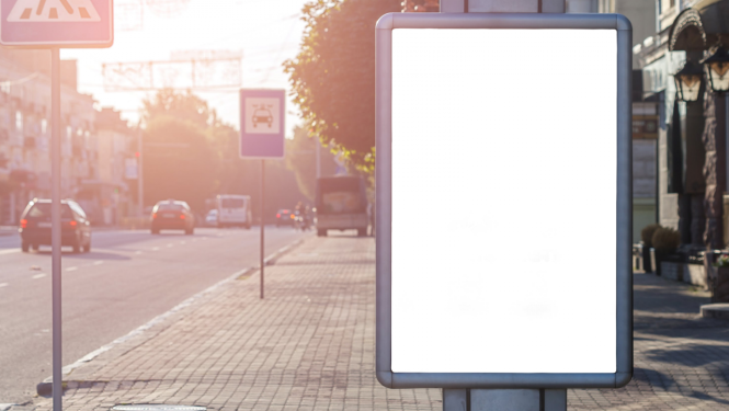 Iela, redzamas gājēju pārejas un pieturas ceļa zīmes. Attēla centrālais objekts - tukšs vides reklāmas stends.