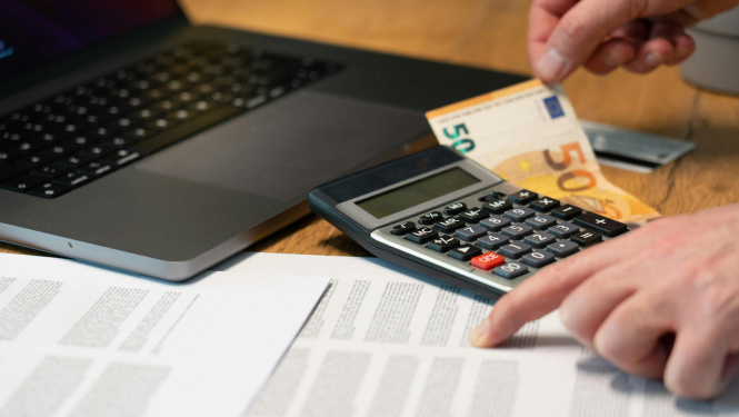 Tuvplānā redzamas rokas pie galda ar dokumentiem, kalkulatoru un klēpjdatoru; viena roka tur 50 eiro banknoti, bet otra norāda uz papīros redzamu informāciju.