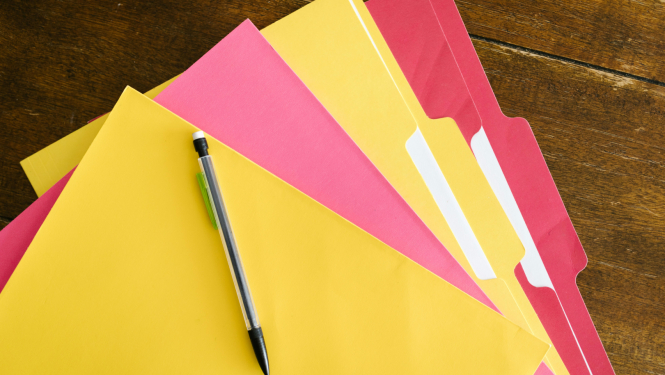 Several yellow and pink file folders are spread across a wooden table, with a black pen placed on top.