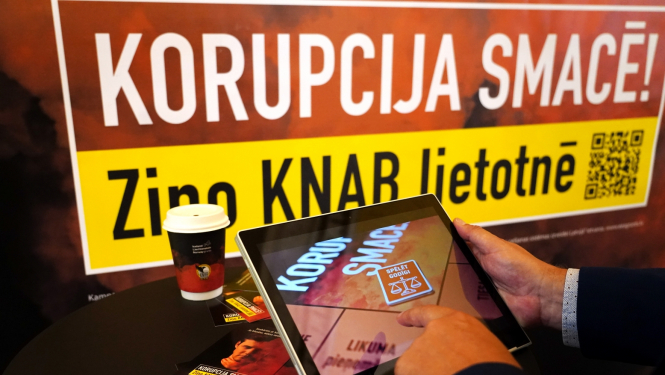 Baneris ar kampaņas devīzi "Korupcija smacē! Ziņo KNAB lietotnē", kā arī attēls ar planšetdatoru, kurā spēlēts kampaņas interaktīvais rīks