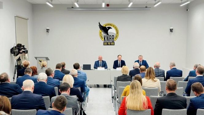 Gaiša zāle, fonā KNAB logo. Telpā ar muguru sēž dažādi cilvēki, zāles galā pie galda trīs vīrieši. No kreisās: KNAB priekšnieks Jēkabs Straume, Eiropas prokurors Gatis Doniks un Latvijas pašvaldību savienības priekšsēdis Gints Kaminskis. 
