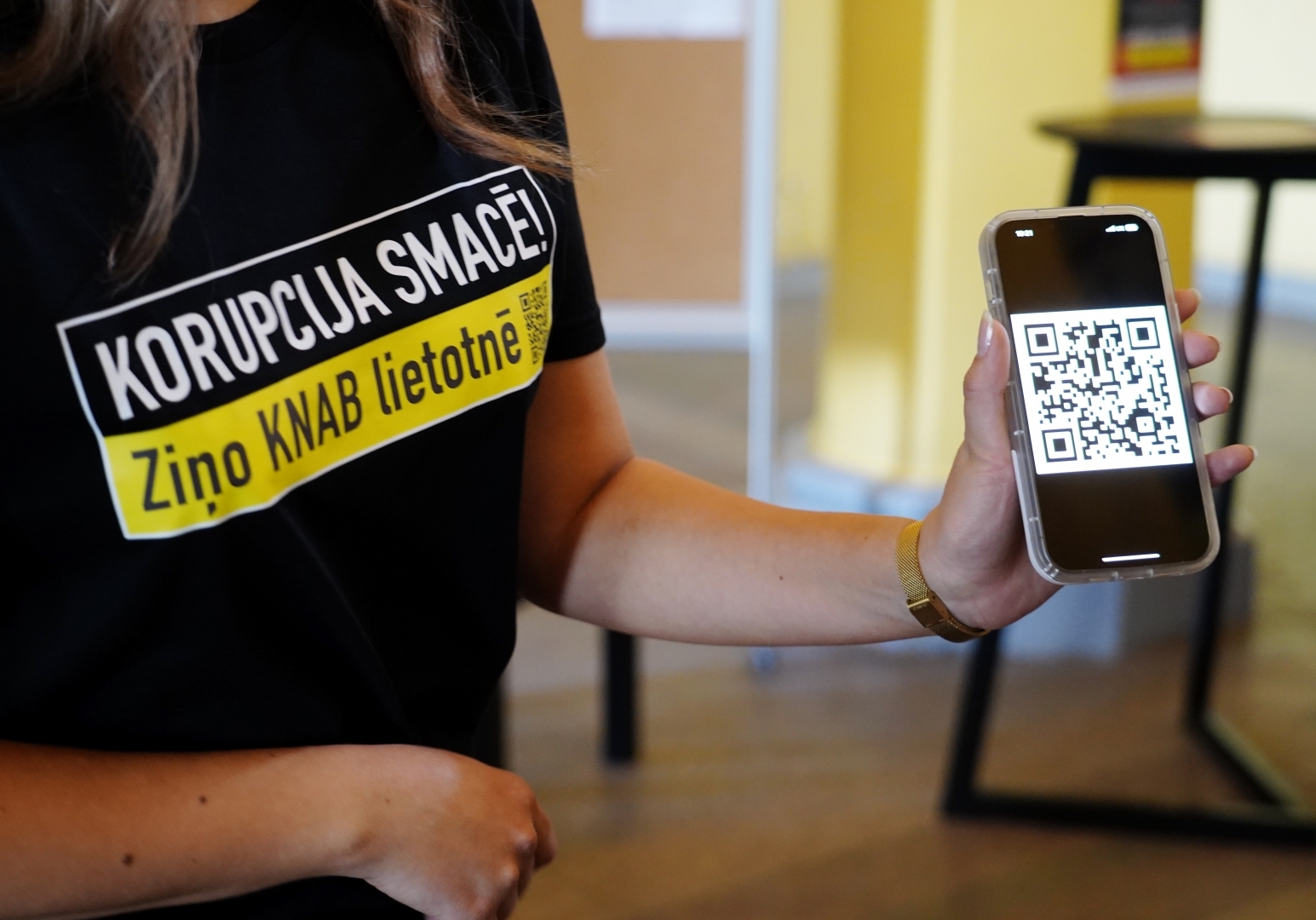 Sieviete ar t-kreklu ar KNAB kampaņas saukli "Korupcija SMACĒ! Ziņo KNAB lietotnē" un telefons ar QR kodu