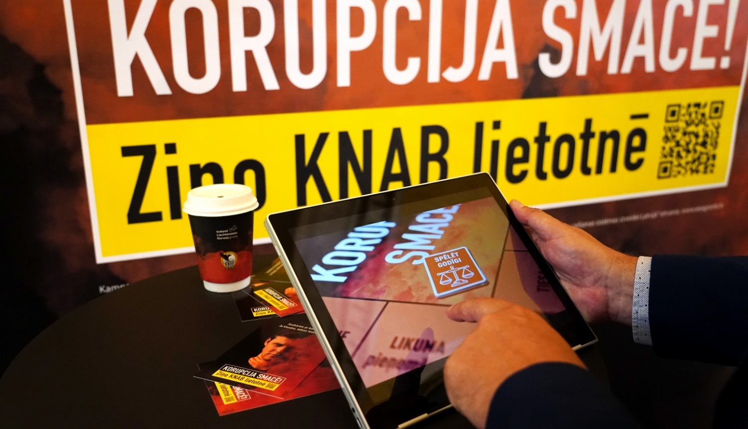 Baneris ar kampaņas devīzi "Korupcija smacē! Ziņo KNAB lietotnē", kā arī attēls ar planšetdatoru, kurā spēlēts kampaņas interaktīvais rīks
