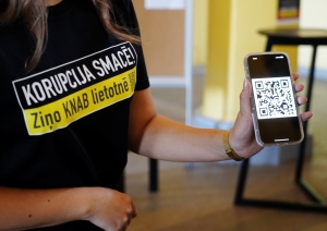 Sieviete ar t-kreklu ar KNAB kampaņas saukli "Korupcija SMACĒ! Ziņo KNAB lietotnē" un telefons ar QR kodu